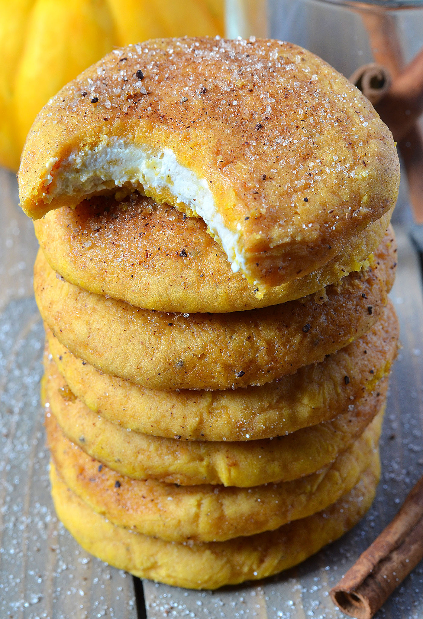 Stack of soft Snickerdoodle Cream Cheese Pumpkin Cookies topped with cinnamon sugar, with a bite revealing the creamy filling inside one cookie. The vibrant pumpkin color and warm spices make them the perfect fall treat.