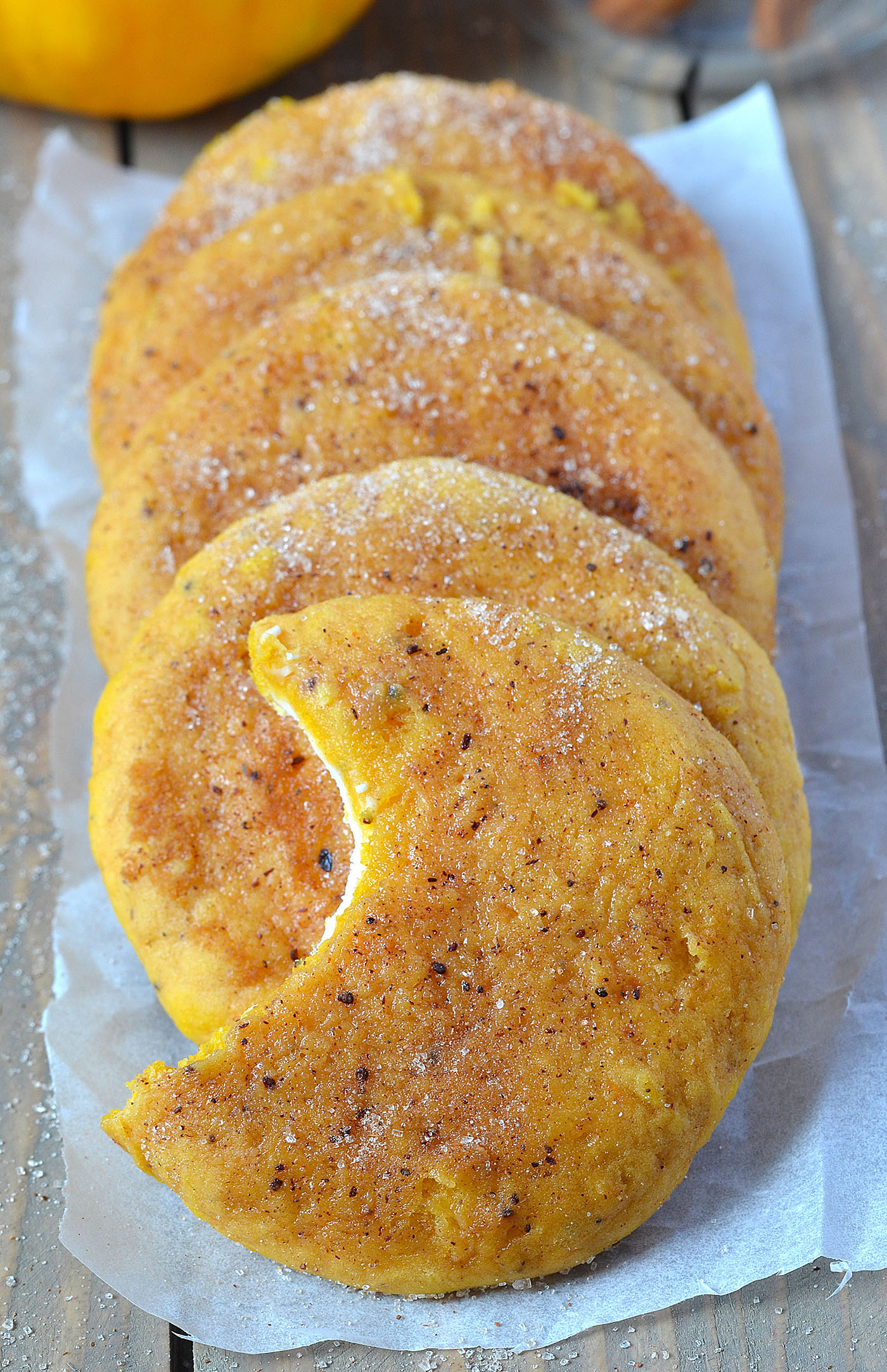 Row of Snickerdoodle Cream Cheese Pumpkin Cookies arranged on parchment paper, each cookie golden brown with cinnamon sugar coating and one cookie showing a bite revealing the creamy filling inside.