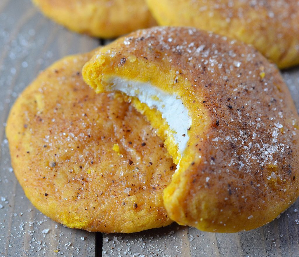 Close-up of a Snickerdoodle Pumpkin Cookie with a bite taken out, showing the creamy filling inside. The cookie is dusted with cinnamon sugar, highlighting its soft, spiced texture.