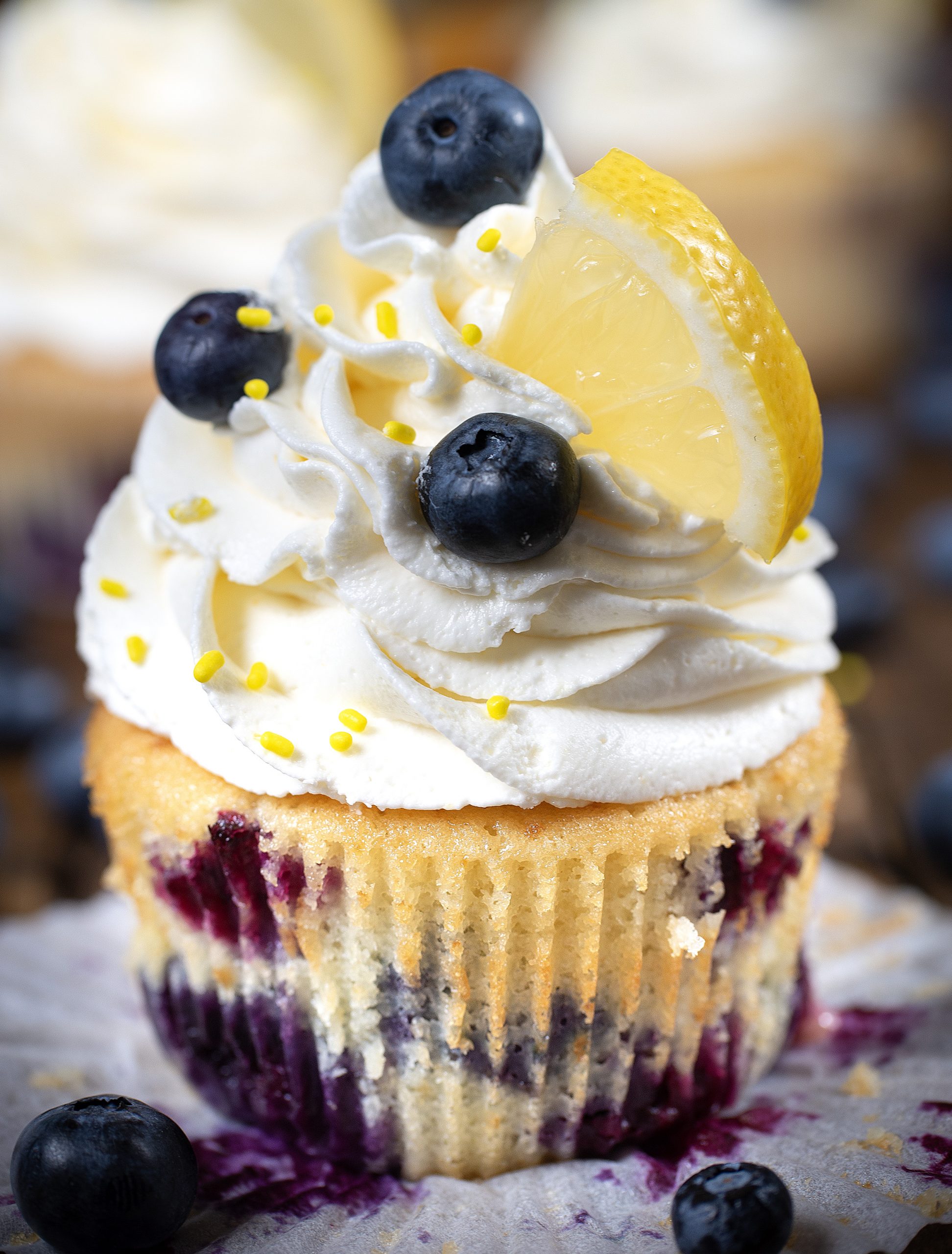 Close-up of a lemon blueberry cupcake topped with a swirl of white frosting, fresh blueberries, and a small wedge of lemon.