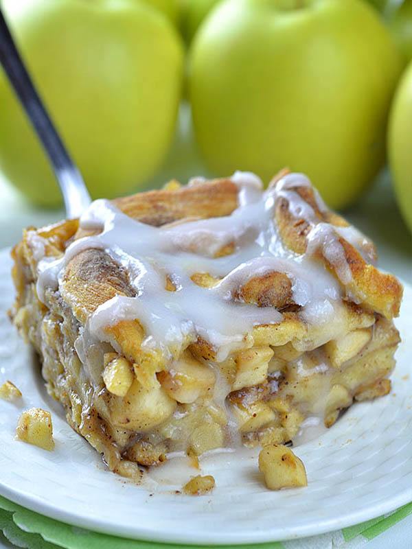 Piece of Caramel Apple Cinnamon Roll Lasagna on a white plate in front of bunch of apples.