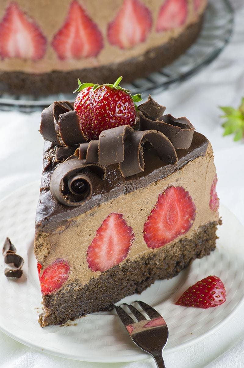 Piece of strawberry chocolate cake on a white plate. Chocolate layer with strawberries inside, garnished with chocolate curls and strawberries on top.
