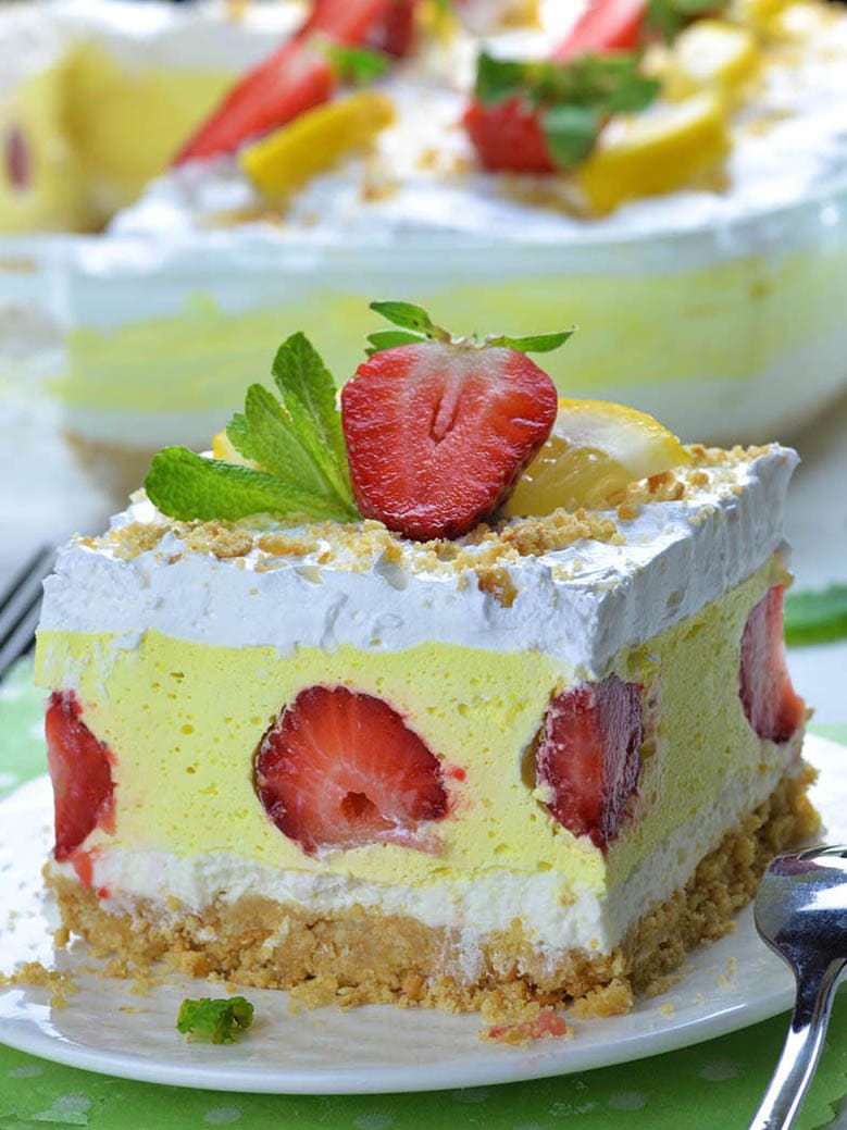 Big piece of Strawberry Lemonade Lasagna on a plate in front of casserole. Yo can see 4 gorgeous layers: bottom Golden Oreo layer, cream cheese layer, yellow lemonade layer with half cut strawberries and top whipped cream layer garnished with strawberries.