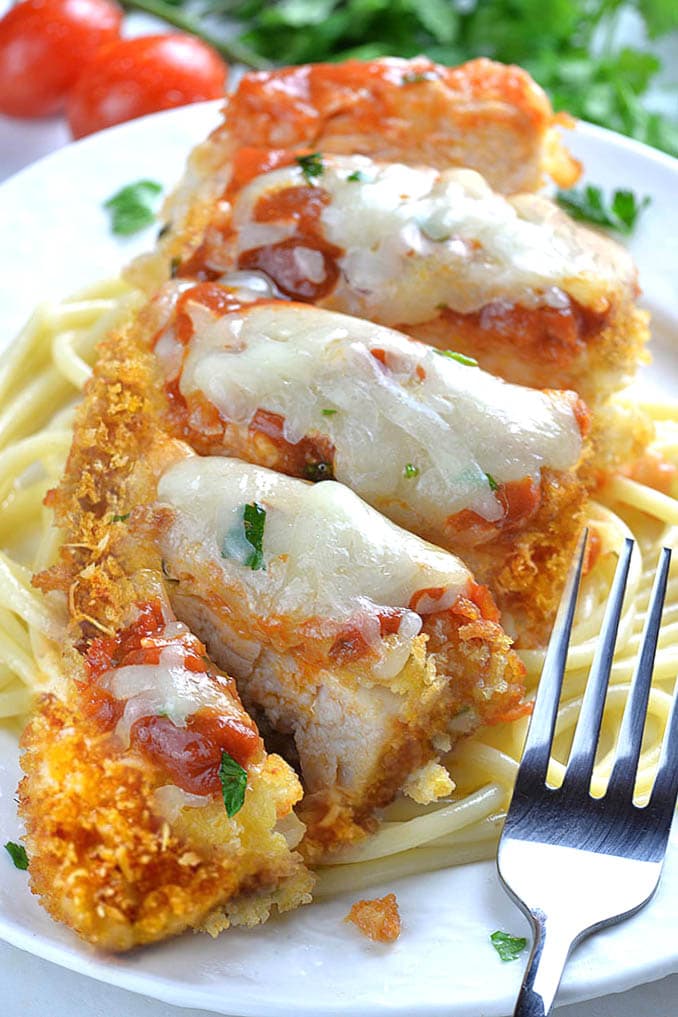Whole piece of Baked Chicken Parmesan on a white plate laying on spaghetti with fork.