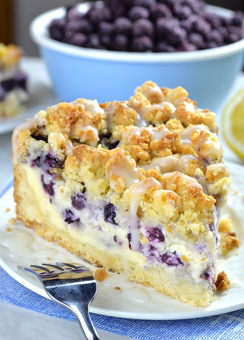 Piece of Blueberry Cheesecake Crumb Cake on a white plate in front of bowl full of fresh blueberries.