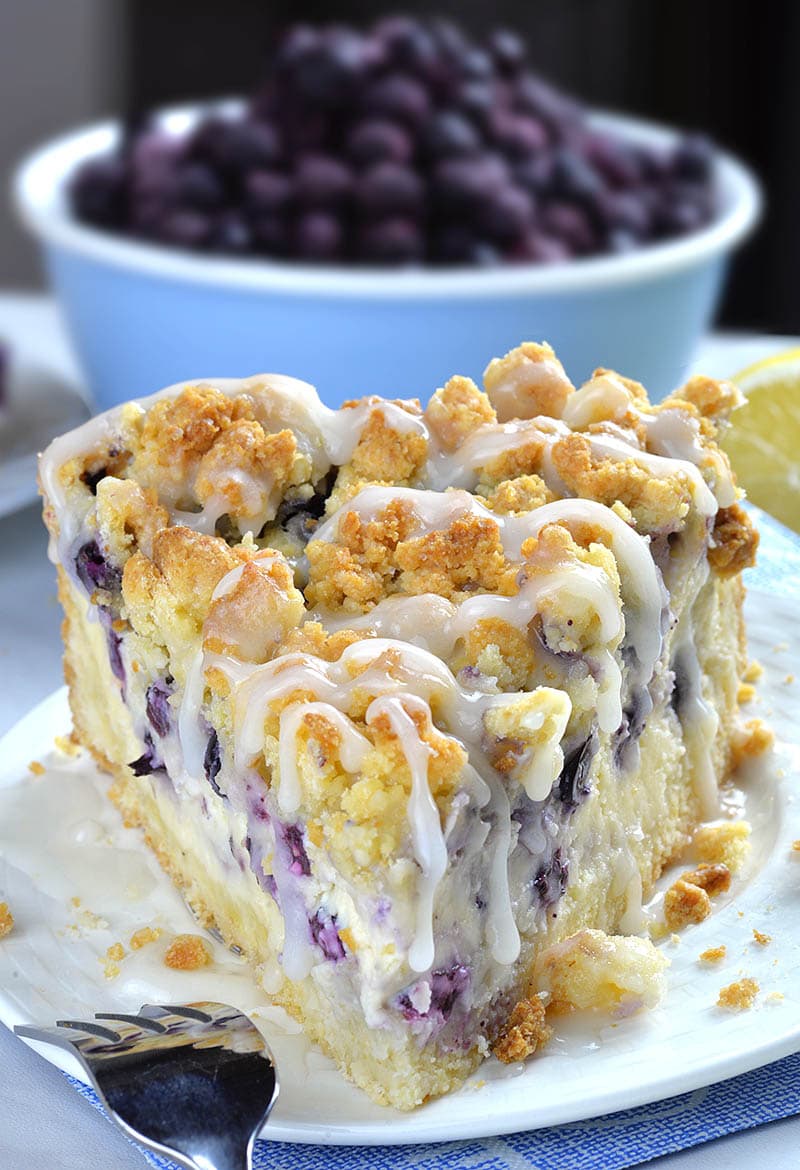 Piece of Blueberry Cheesecake Crumb Cake on a white plate in front of bowl full of fresh blueberries.