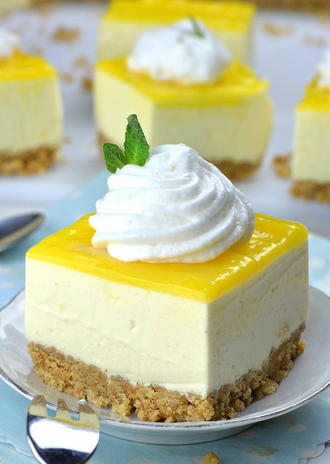 Piece of No Bake Lemon Cheesecake Bar on a white plate with cool whip topping in front of couple of pieces.