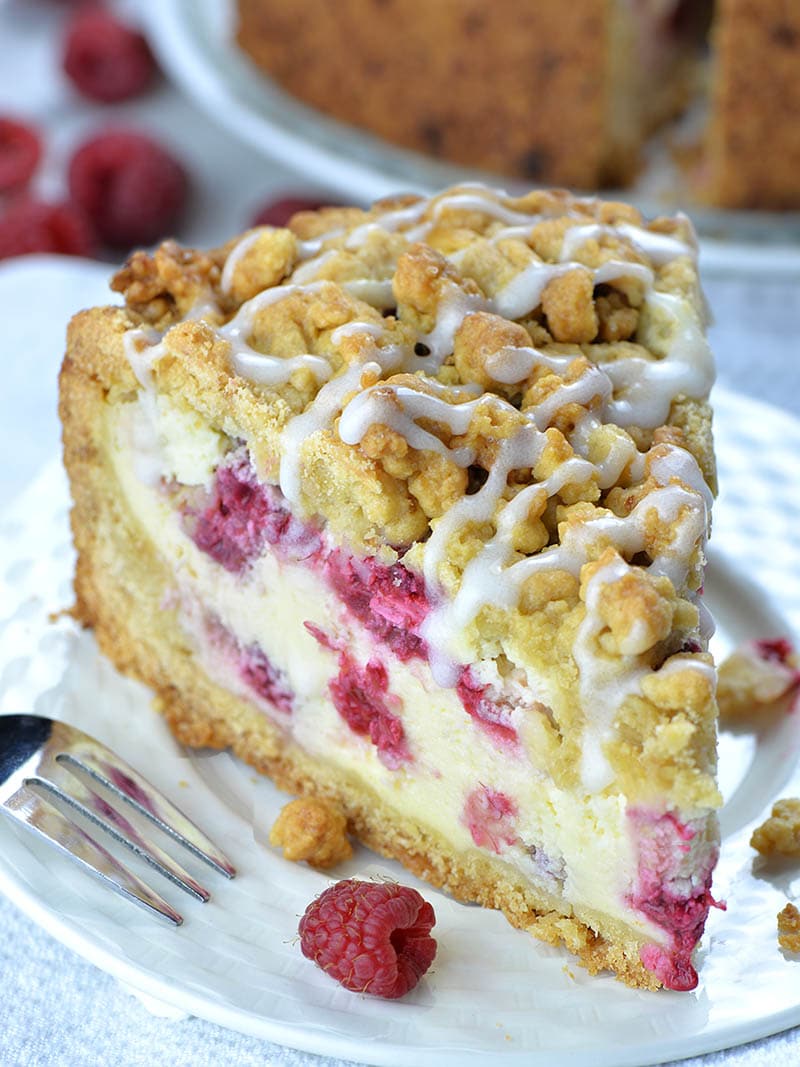 Big piece of Raspberry Cheesecake Crumb Cake on white plate with couple of raspberries beside. 