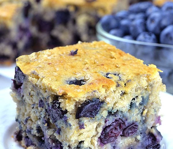 Piece of Blueberry Breakfast Cake on a white plate with a bunch of cakes and blueberries behind.