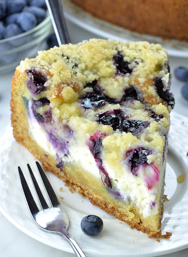 Piece of blueberry coffee cake served with a fork and fresh blueberry on white plate.