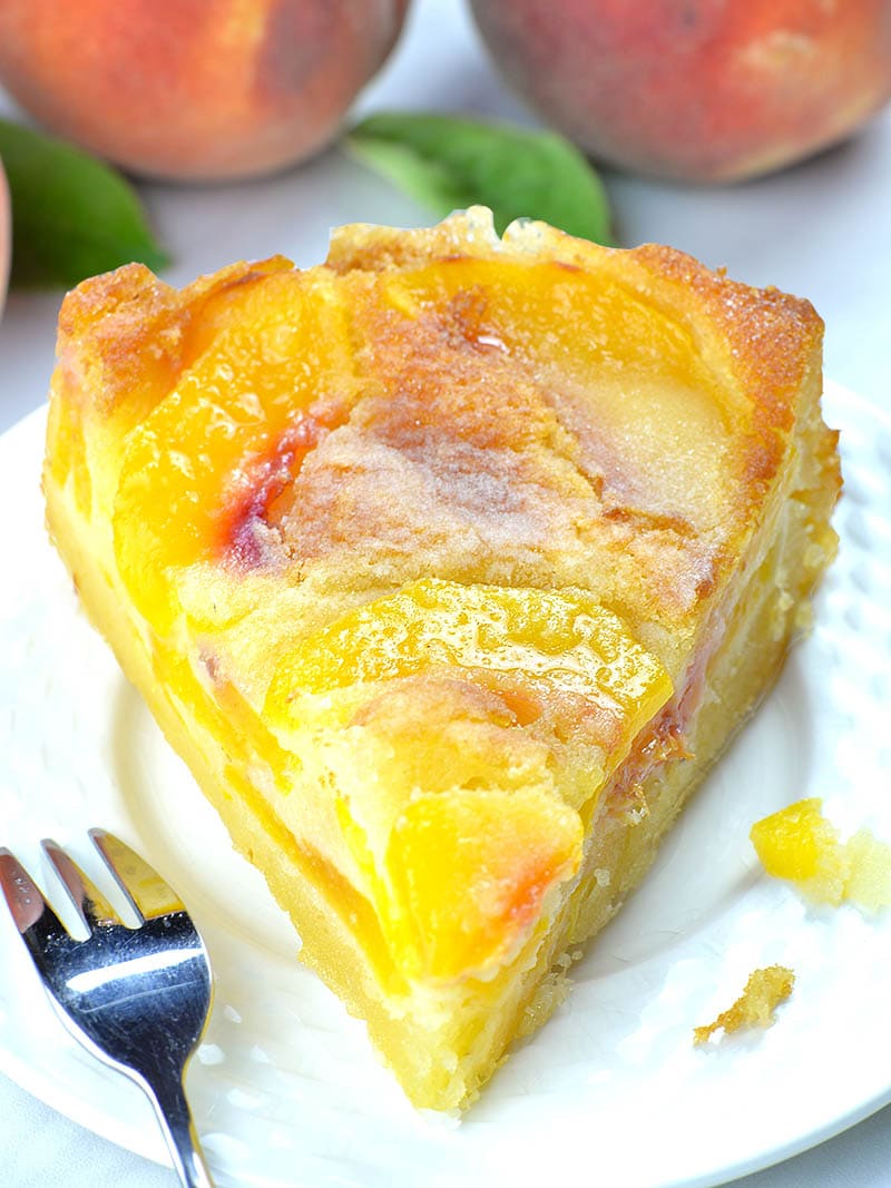 Piece of peach cake on white plate - bottom part is packed with peaches and has a dense, custard texture, while the top layer has cake-like texture.
