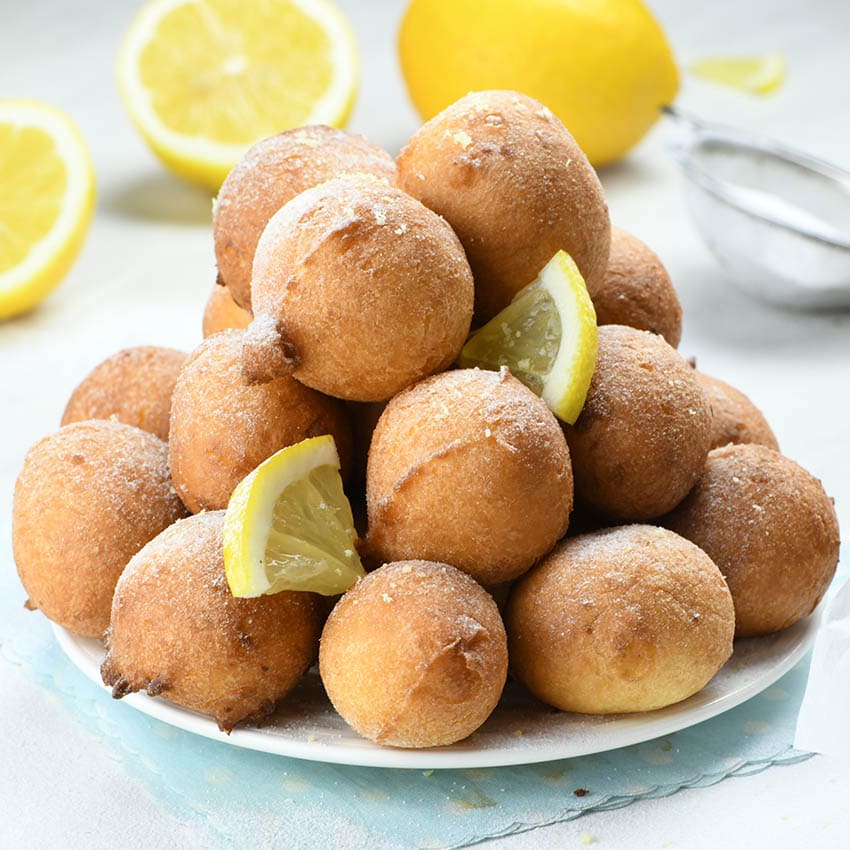 Lemon Fritters on a white plate garnished with two lemon pieces with couple of lemons behind.