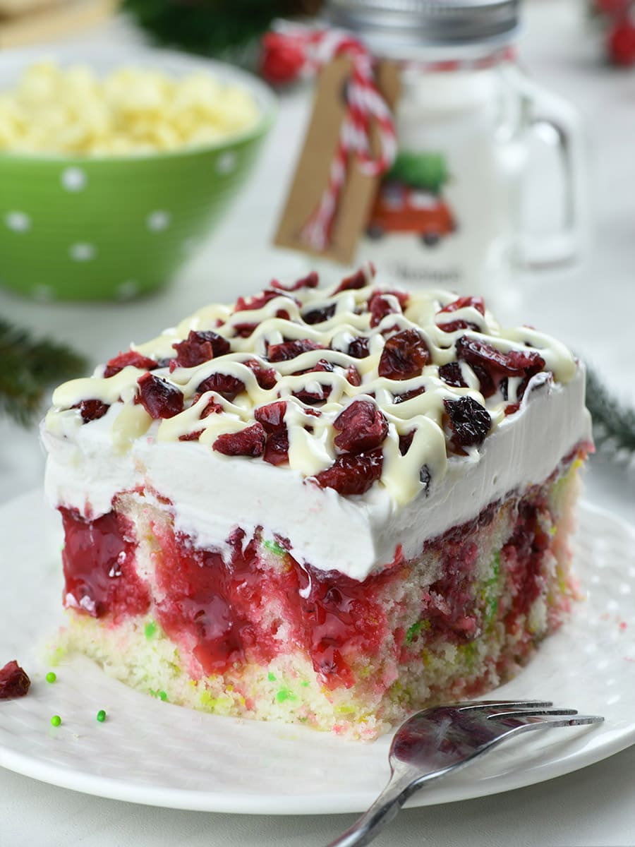 Cranberry Poke Cake on a white plate with Christmas decorations behind.