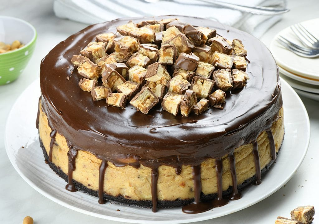 Whole Peanut Butter Cheesecake on a white plate garnished with chocolate ganache and Snicker bar chunks.
