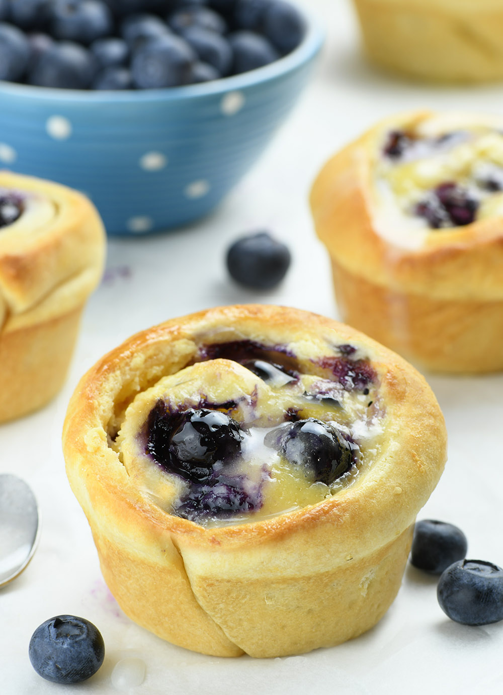 Blueberry Crescent Roll Muffins on a table with couple of muffins behind and garnished with fresh blueberries.