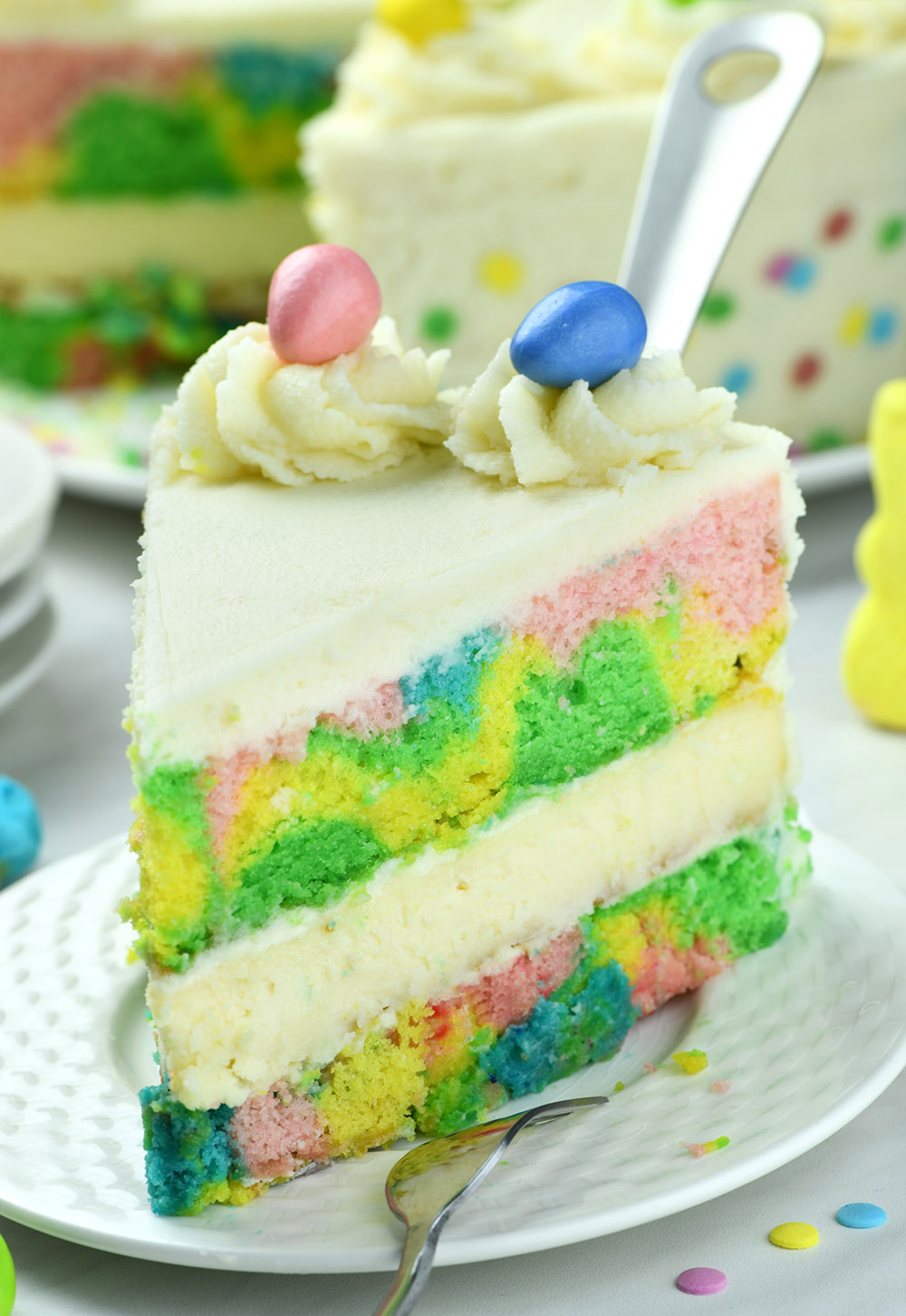 Smooth and creamy white chocolate cheesecake hidden between colourful cake layers and white chocolate buttercream on a white plate , decorated with frosting and Easter eggs.
