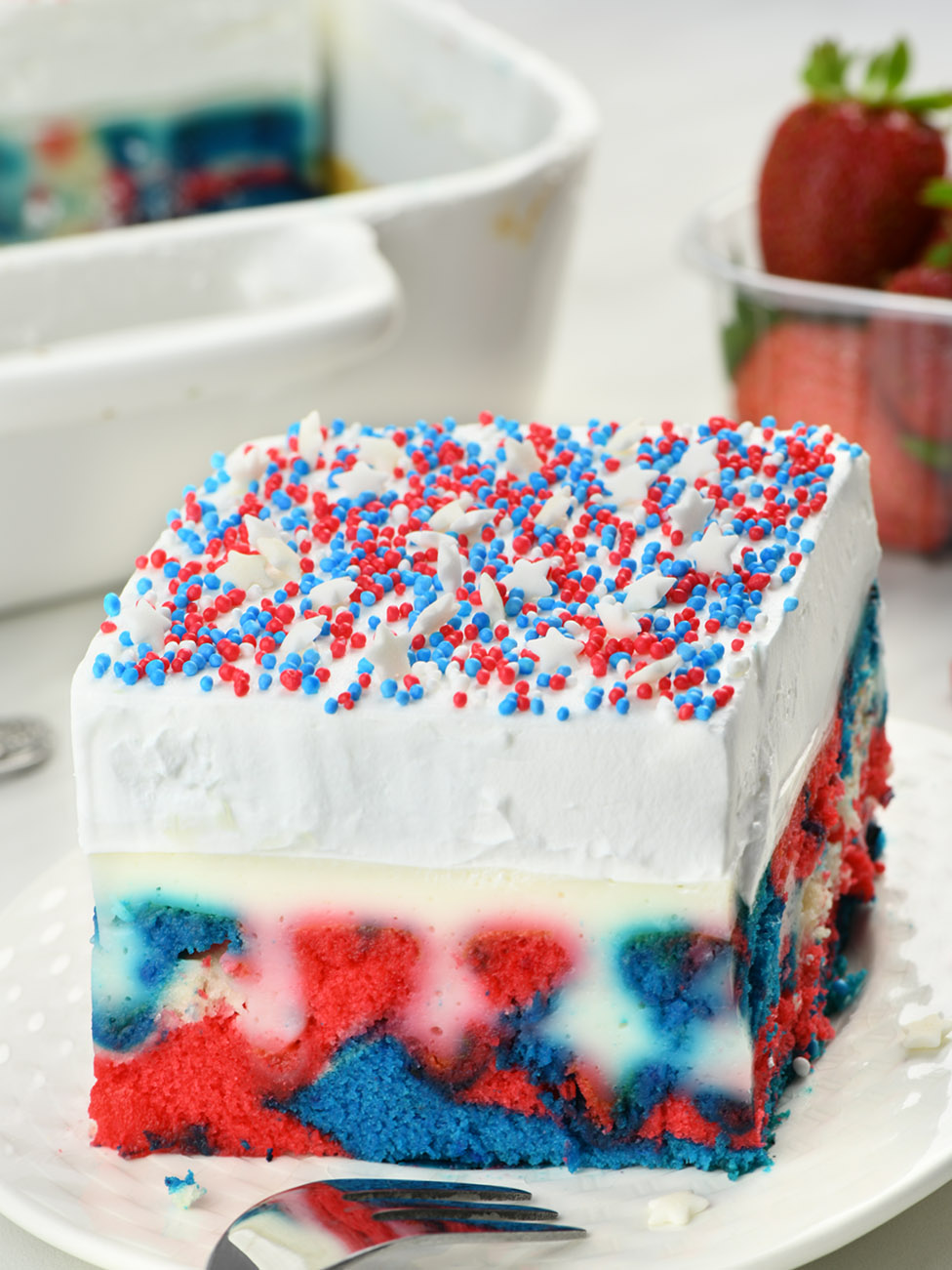 Red, white and blue Patriotic Poke Cake,  on a white plate garnished with red, white and blue funfetti,