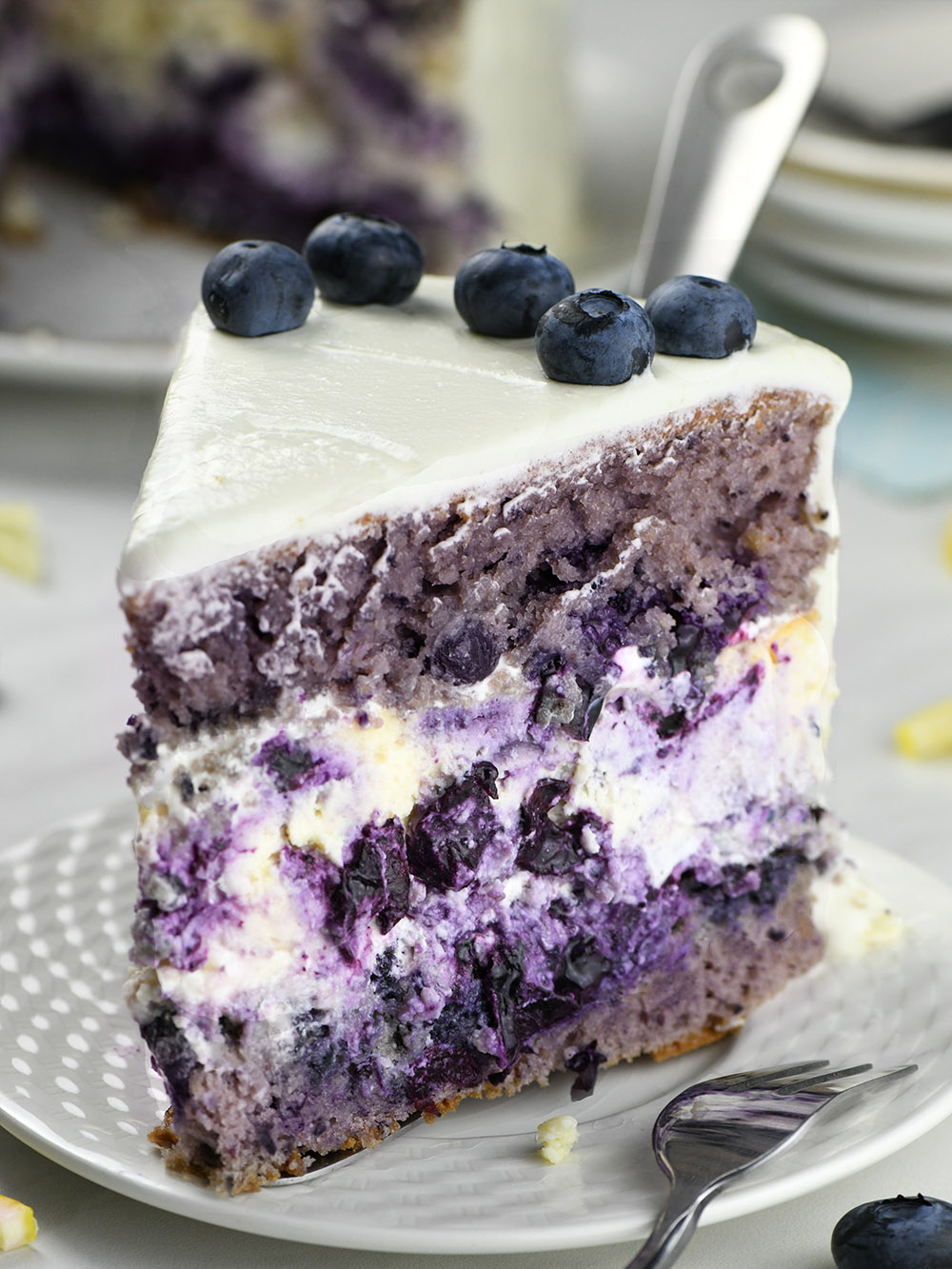 Piece of layered Blueberry Cheesecake Cake on a white plate with a whole cake behind.