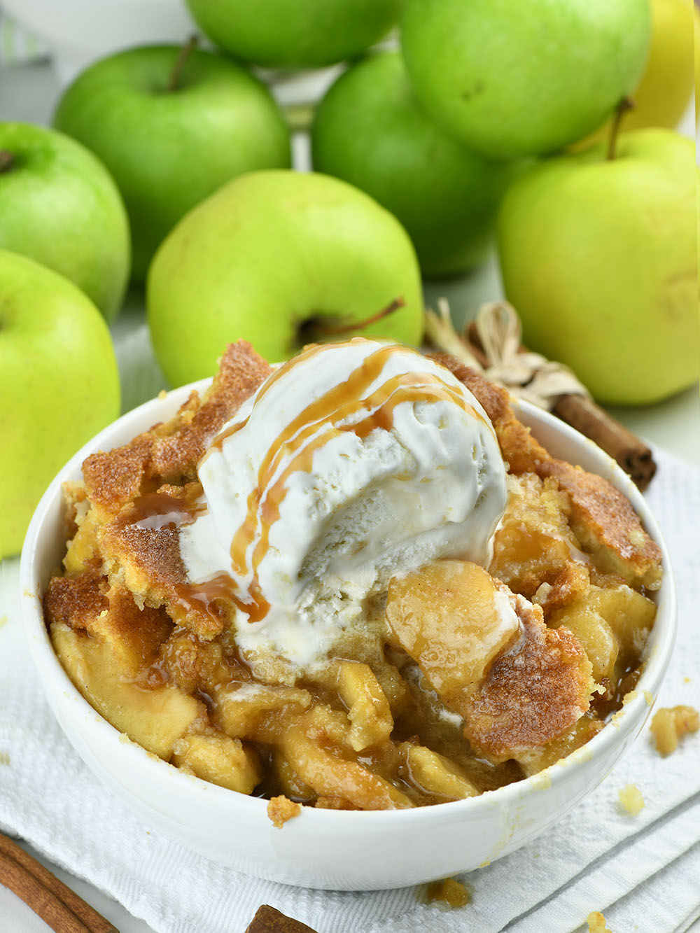Apple Cobbler in a white bowl decorated with a large scoop of ice cream, in front of a pile of green apples.