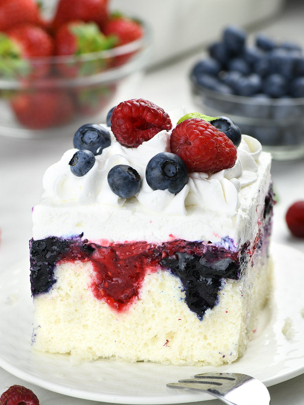 Summer berry poke cake on a white plate, garnished with berries and topped with cool whip.