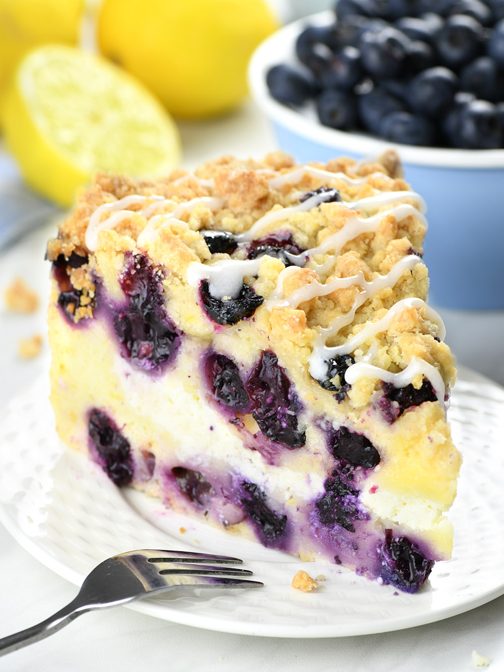 A delectable piece of Lemon Blueberry Coffee Cake is presented on a clean, white plate. The cake has a moist, golden-brown crumb texture, with dots of bright, juicy blueberries and a delicate lemon glaze. Fresh lemons and a scattering of ripe blueberries in the background provide a colorful contrast. The composition is shot from a side angle, emphasizing the fluffy thickness of the cake slice.