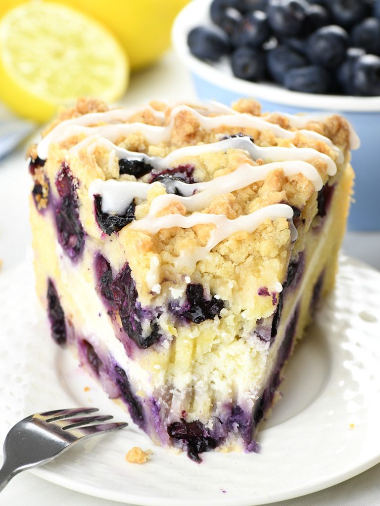 Front view of a piece of Lemon Blueberry Coffee Cake on a white plate, cut into with a fork. The cake has a golden crumb texture and a generous sprinkling of blueberries throughout, topped with a zesty lemon glaze. In the blurred background, a fresh lemon and a handful of ripe blueberries are present, signifying the key ingredients.