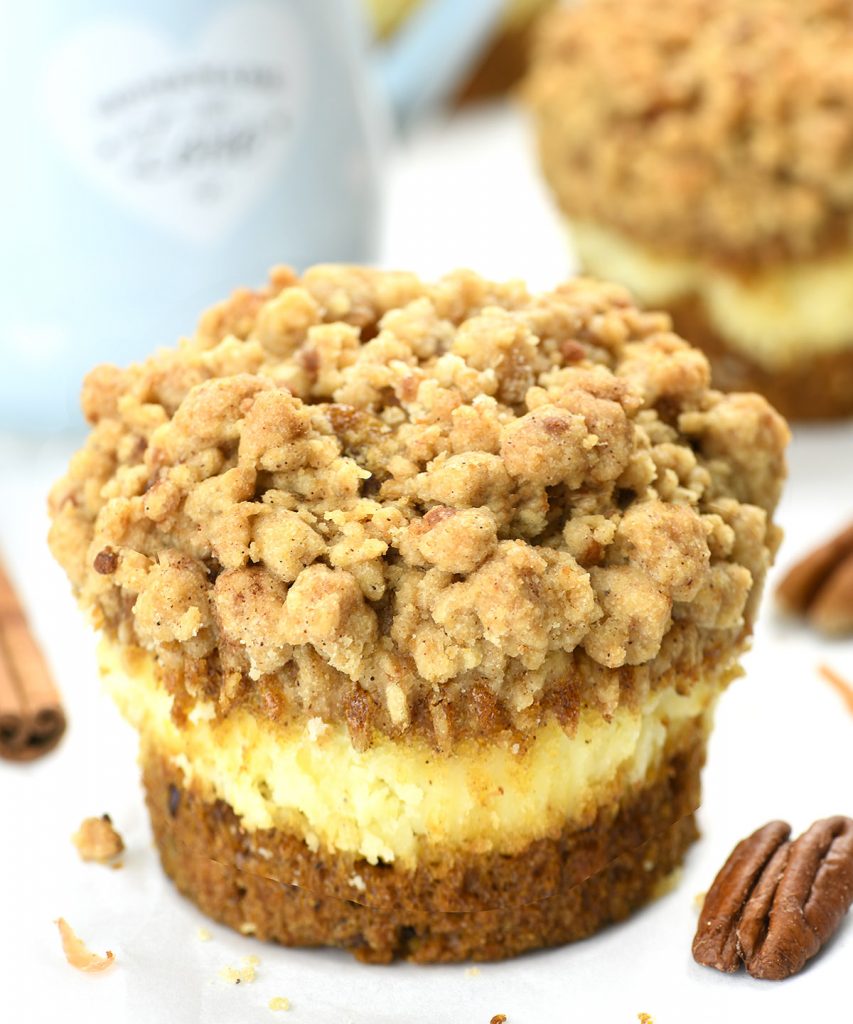 Close-up of a mini carrot cake cheesecake with a crumbly streusel topping, garnished with pecans, against a blurred background featuring a blue mug and additional cheesecakes.