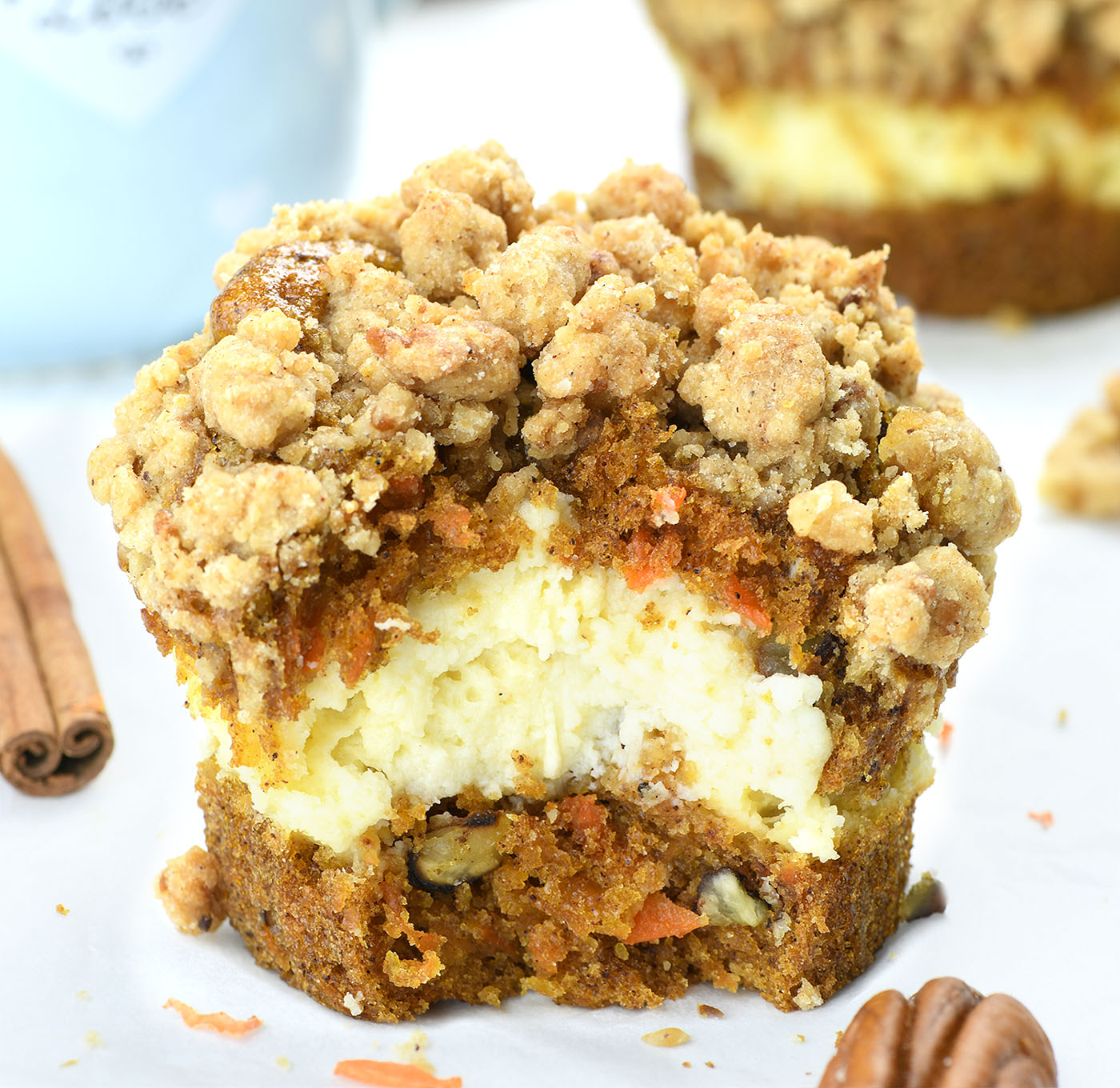 Mini carrot cake cheesecake with a streusel topping and pecan pieces, displayed on a white surface with additional cakes and a blue mug in the background.