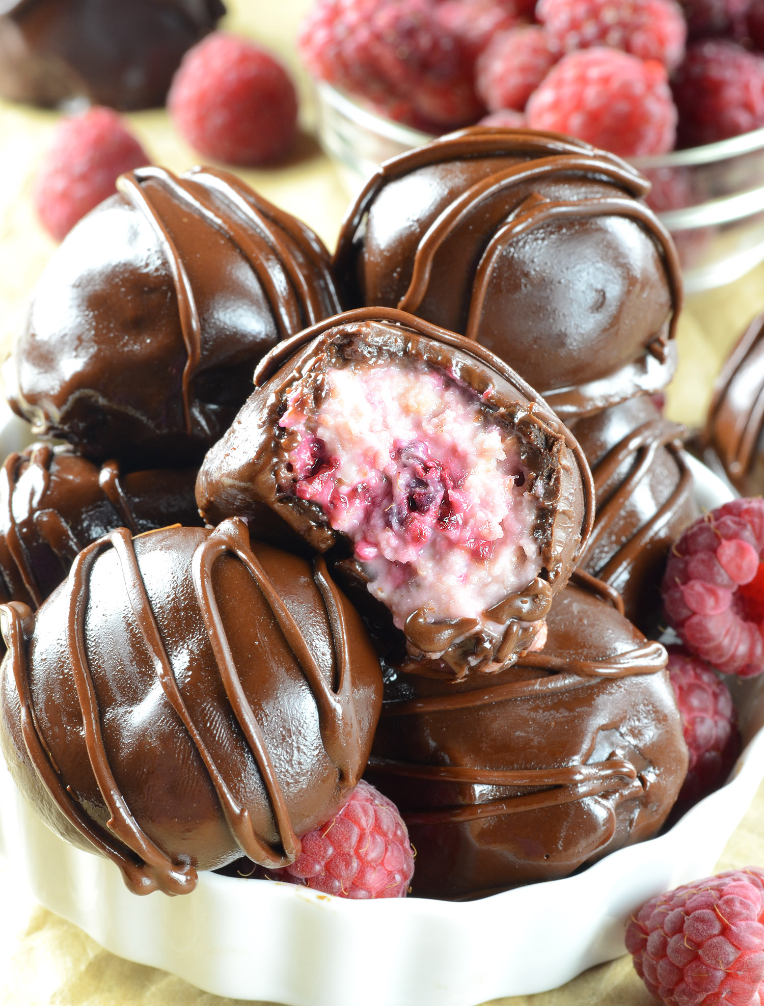 Decadent raspberry truffles coated in dark chocolate with a rich raspberry filling, drizzled with melted chocolate, presented in a white bowl with fresh raspberries in the background.