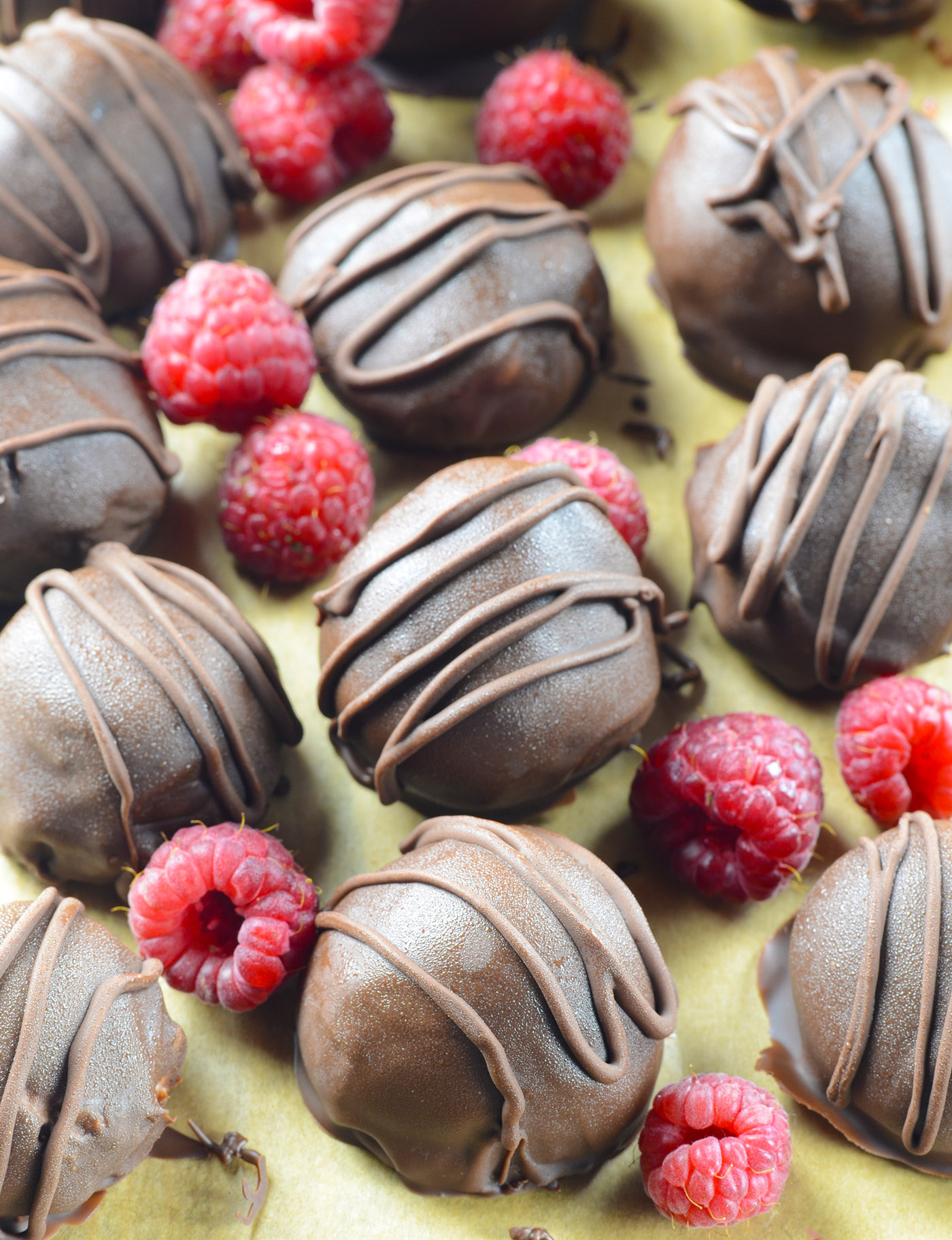 Assorted chocolate truffles with elegant chocolate drizzle, paired with vibrant fresh raspberries on a parchment-covered surface.