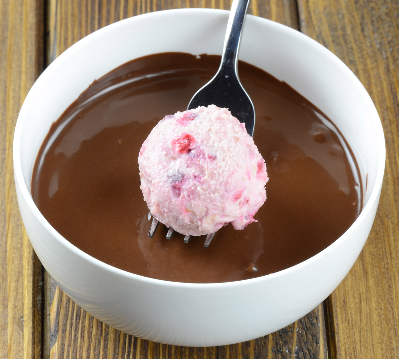 A raspberry truffle ball  dipped into a bowl of smooth melted chocolate, held by a fork, ready for coating.