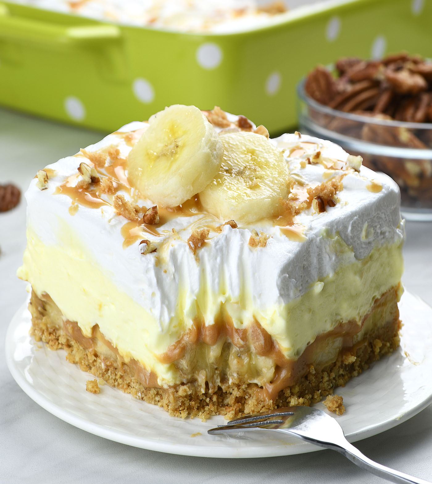 A slice of banoffee pie lasagna on a plate, showing layers of biscuit crust, caramel, banana, and cream, topped with banana slices and drizzled caramel, with nuts sprinkled on top.