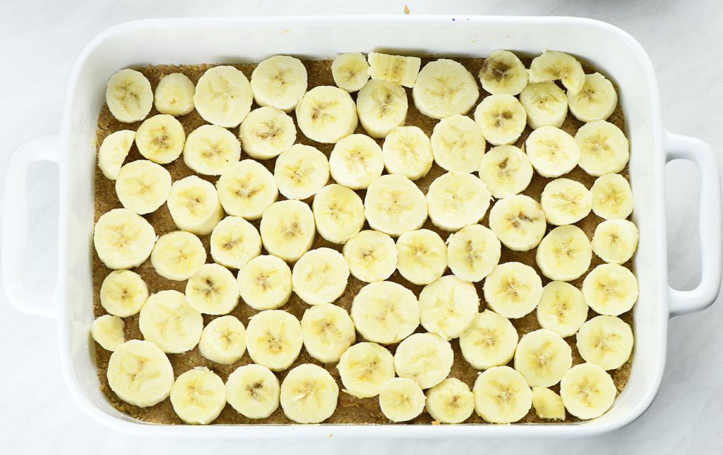 A graham cracker crust in a white baking dish topped evenly with sliced bananas, ready for the next layer of the dessert.