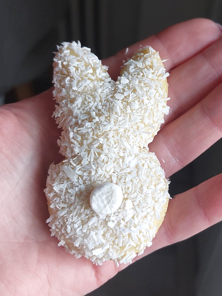 The image shows a close-up of a hand holding a small, Easter bunny-shaped cookie covered in shredded coconut, with a single round decoration in the center representing the bunny's tail. 