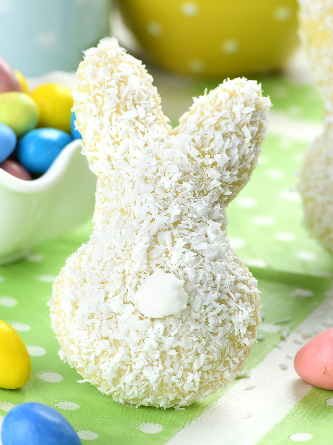 An Easter bunny-shaped cookie coated with shredded coconut to resemble fur, standing upright on a table with a polka-dotted green tablecloth. The background suggests a festive Easter setting with pastel-colored Easter eggs in soft focus.