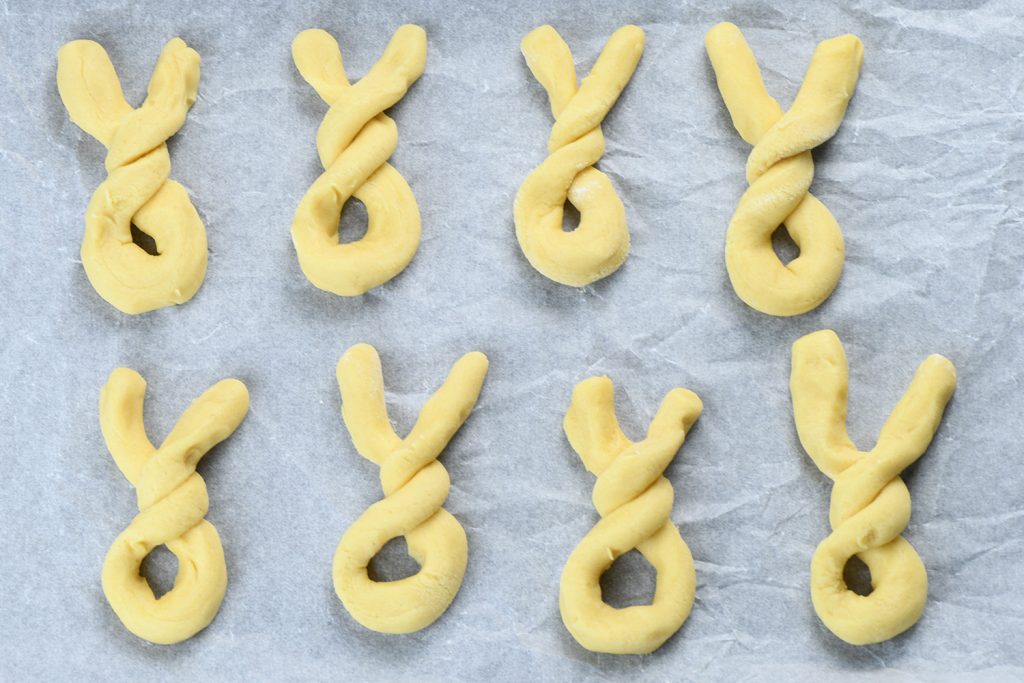 Raw cookie dough shaped into Easter bunny figures before baking, laid out on parchment paper. The dough is twisted to form bunny ears atop round bodies.