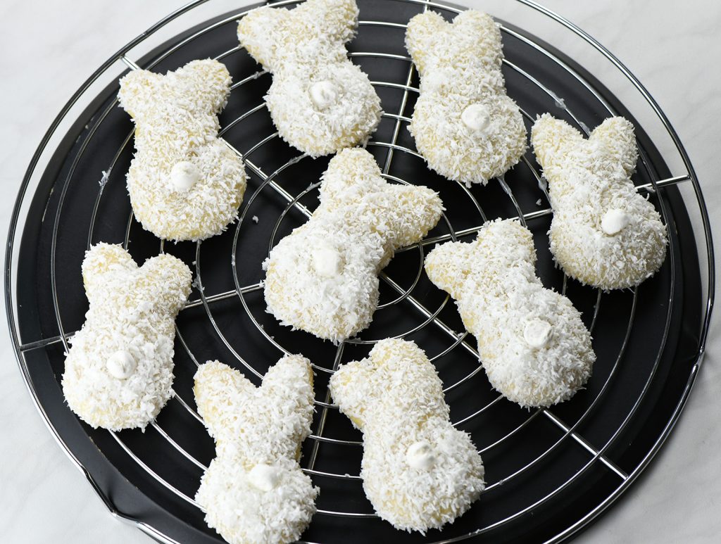 Easter bunny-shaped cookies on a cooling rack. Each cookie is coated with shredded coconut to give the appearance of soft, white fur, and has a small dollop of white icing to represent the tail. 