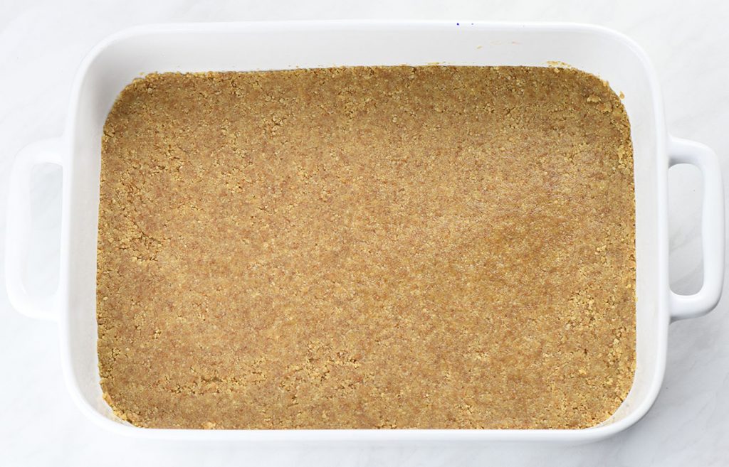 Graham cracker crumbs pressed into an even layer in a white baking dish, forming a crust base for a dessert.