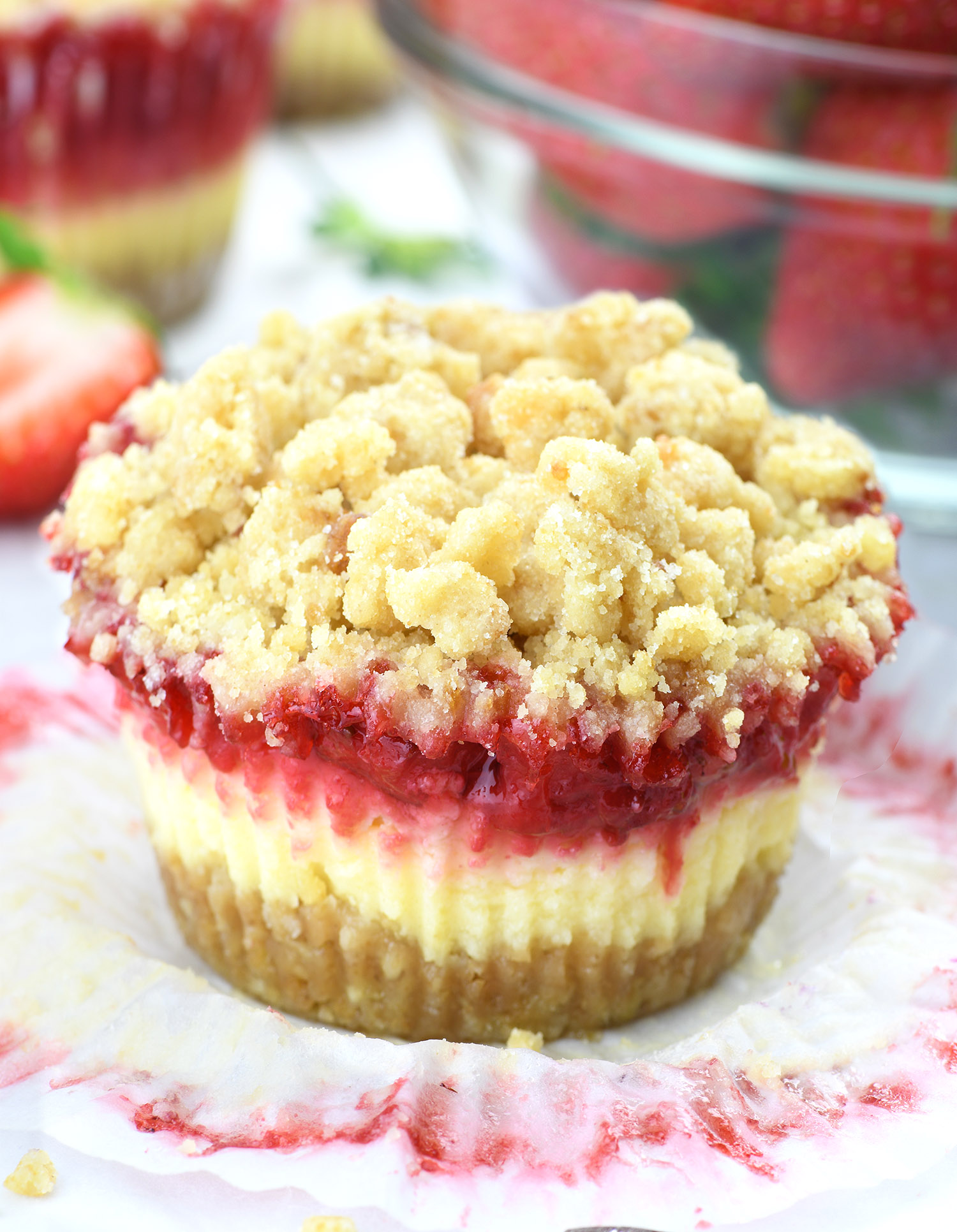 A close-up of a mini strawberry cheesecake with a crumble topping, presented on a piece of parchment paper. The cheesecake has a golden-brown crust at the base, a creamy white cheesecake layer in the middle, and a bright red strawberry layer on top, all covered with a generous amount of light golden crumble. 