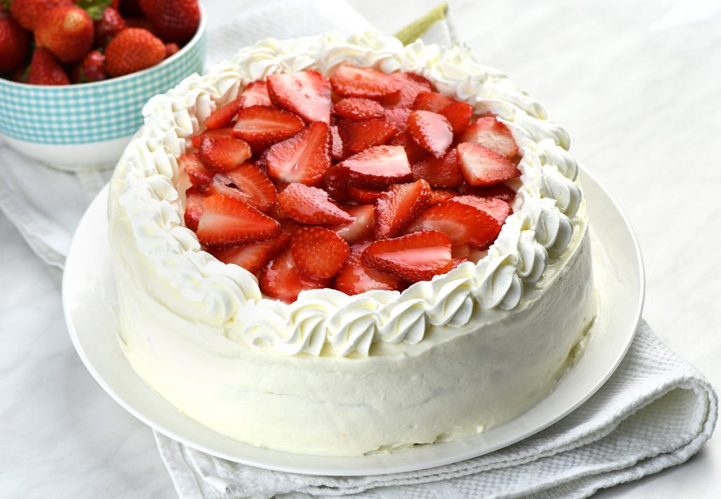 Whole strawberry cake on a plate, beautifully decorated and ready to be served. The cake is covered in a smooth white frosting and bordered with elegant whipped cream swirls around the top edge.
