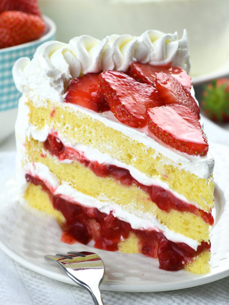 A slice of strawberry cake on a plate. The cake features multiple layers, with a bright, moist yellow sponge, creamy white frosting, and a generous amount of vibrant red strawberry slices and jam between the layers. 