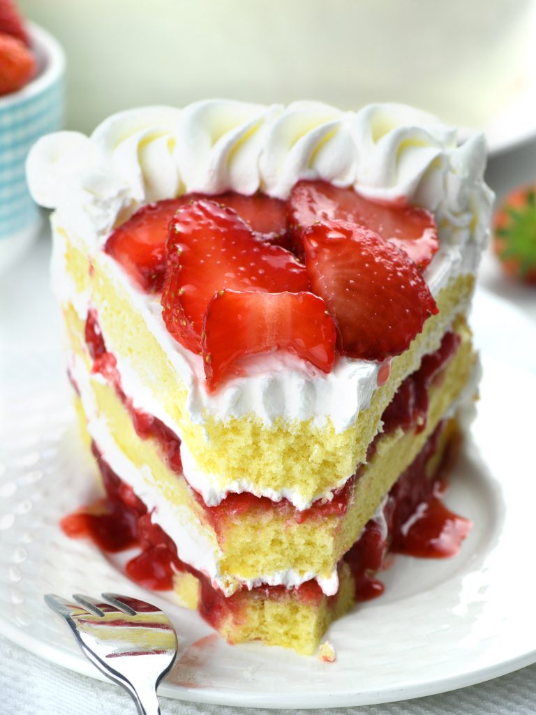 A close-up view of a slice of strawberry cake. This cake features multiple layers of soft, light yellow sponge cake separated by thick layers of creamy white frosting and a generous amount of glossy, deep red strawberry pie filling. 