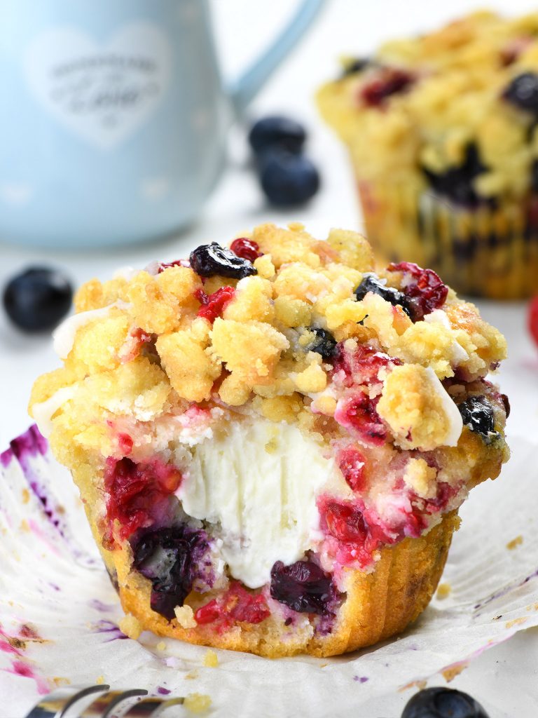 The camera zooms in on a berry cream cheese muffin with a streusel topping that crumbles. It is filled with cream cheese and contains fresh juicy berries like raspberries and blueberries. Some parts of the paper wrapper are peeled back to expose the tender inside part as well as rich colors from the fruits. Behind it, there is a pale blue ceramic coffee cup bearing a heart symbol and more fresh blueberry fruits.