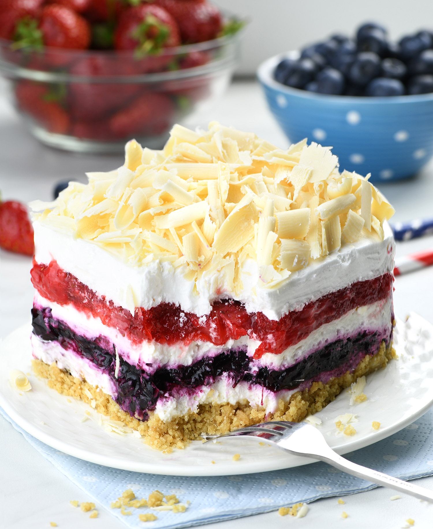 Close-up of a vibrant No Bake Berry Cheesecake Lasagna with layers of creamy cheesecake, red and purple berry fillings, topped with white chocolate shavings. Fresh strawberries and blueberries in bowls are in the background.