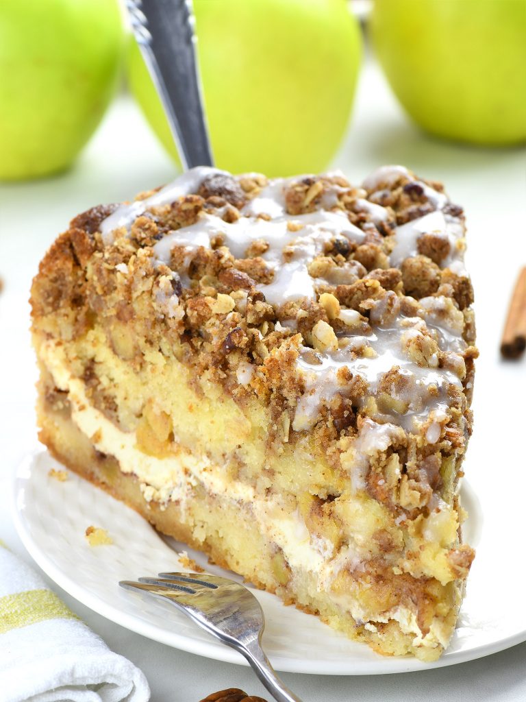 Close-up of a slice of apple coffee cake with a crumbly streusel topping and a drizzle of icing, placed on a white plate with fresh green apples in the background.