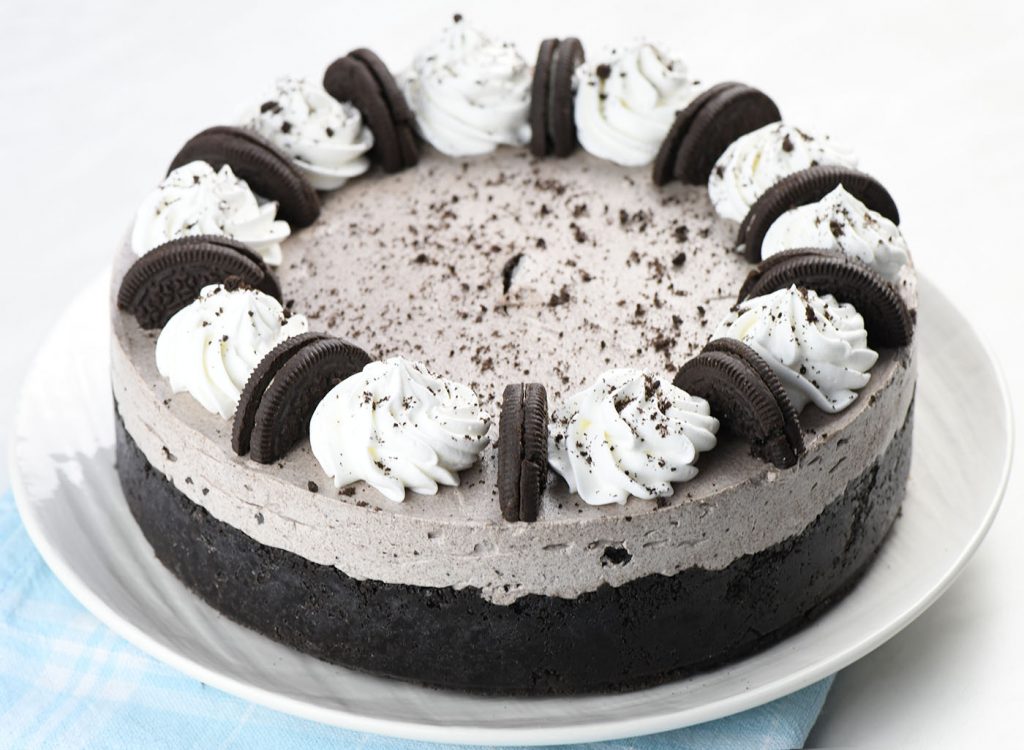 A close-up of a No Bake Oreo Cheesecake, featuring a creamy Oreo filling with a dark chocolate crust. The cheesecake is garnished with whipped cream swirls and Oreo cookie halves arranged around the edges, with a sprinkle of crushed Oreo crumbs on top.