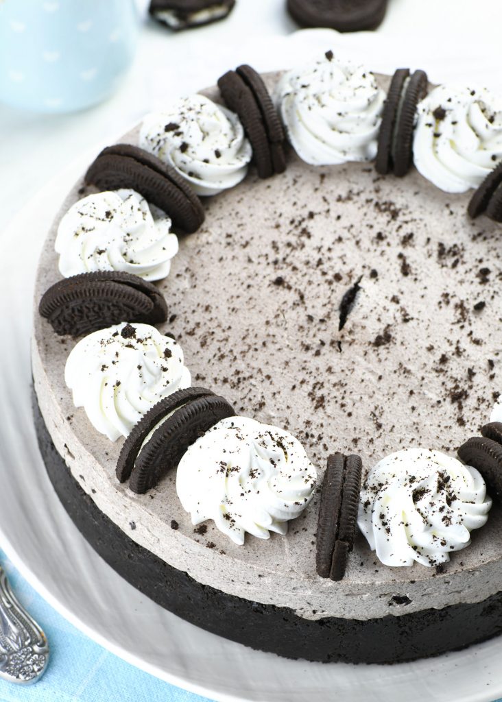 An overhead shot of a No Bake Oreo Cheesecake. The top is decorated with evenly spaced whipped cream swirls and Oreo cookie halves, creating a circular pattern. Oreo crumbs are lightly scattered across the surface of the cheesecake.