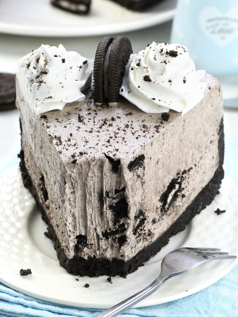 A partially eaten slice of No Bake Oreo Cheesecake. The layers of the Oreo cookie crust, creamy filling with cookie chunks, and whipped cream topping are clearly visible. The slice is served on a white plate with a fork.