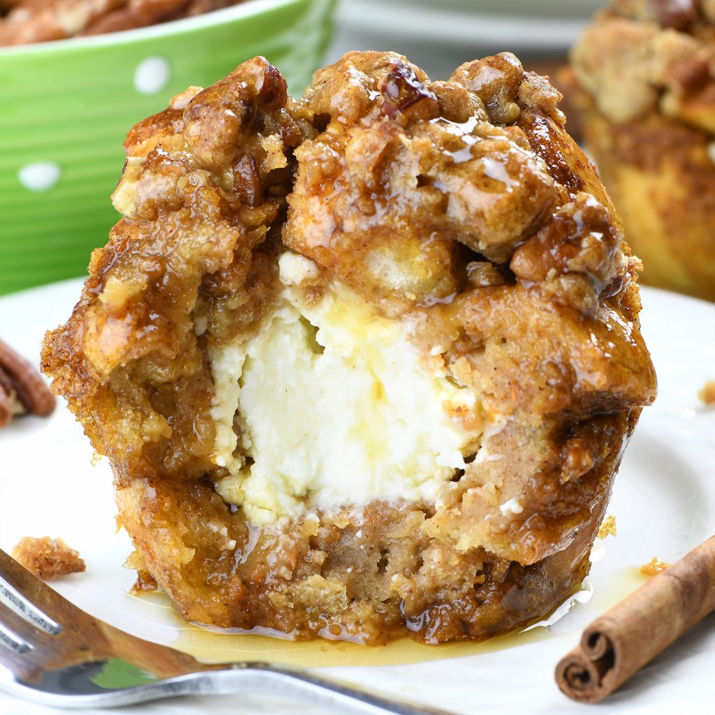 A Pumpkin French Toast Muffin with a bite taken out, revealing a creamy, custard-like center. The muffin is golden brown, with a crunchy streusel topping and drizzled with syrup. The warm pumpkin spice flavors complement the soft, gooey interior, making this muffin a decadent autumn treat. A cinnamon stick and fork are placed beside the muffin for a cozy presentation.