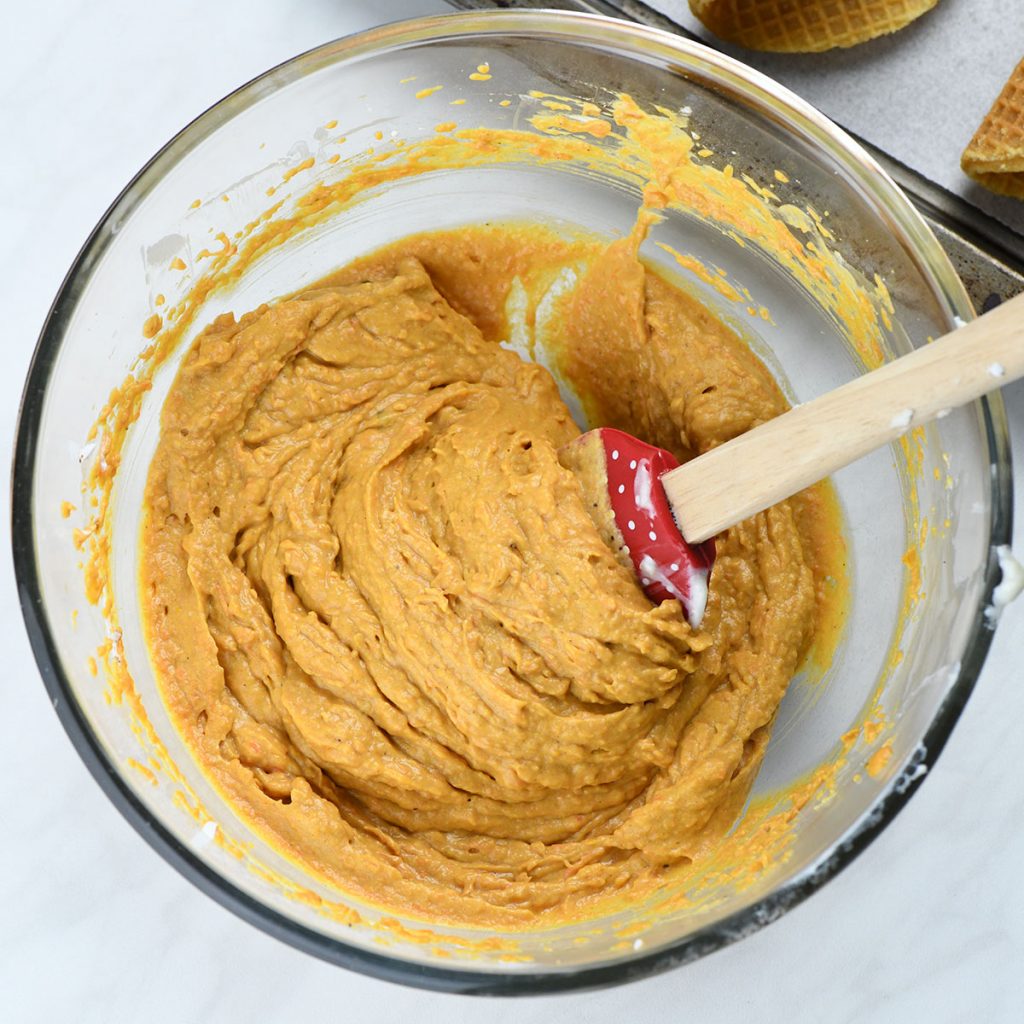 A bowl of smooth pumpkin spice latte mousse being mixed with a red spatula, showing a creamy texture and vibrant orange color.